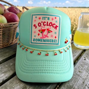 May include: A light teal trucker hat with a decorative cherry chain and the phrase "IT'S 5 O'CLOCK SOMEWHERE!" on the front. The hat has a mesh back and a curved brim. The text is in red and pink.