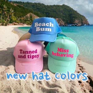 May include: Three trucker hats with different colors and text. The blue hat says "Beach Bum", the pink hat says "Tanned and tipsy", and the green hat says "Miss behaving".