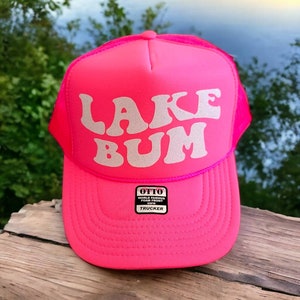 May include: A pink trucker hat with a white sparkly "LAKE BUM" design. The hat has a mesh back and a black label that reads "OTTO WORLD FAMOUS FOAM FRONT TRUCKER".