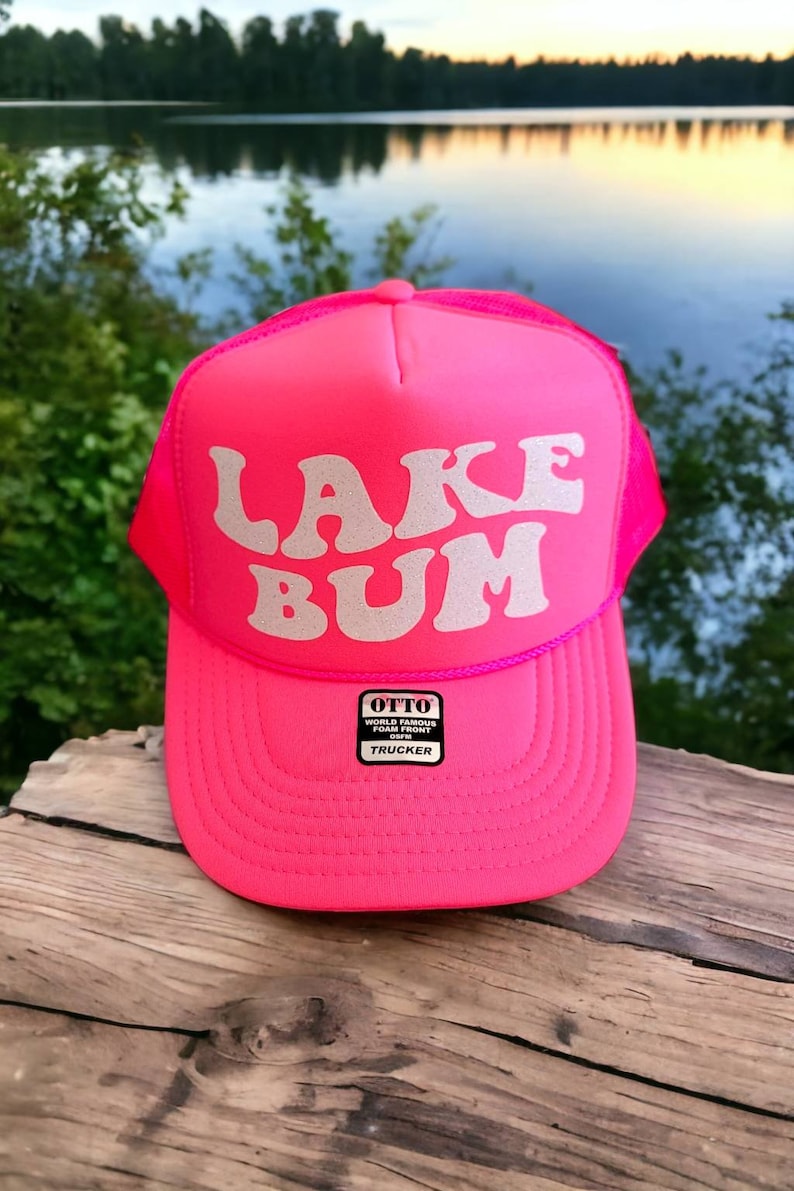 May include: A pink trucker hat with a white sparkly "LAKE BUM" design. The hat has a mesh back and a black label that reads "OTTO WORLD FAMOUS FOAM FRONT TRUCKER".