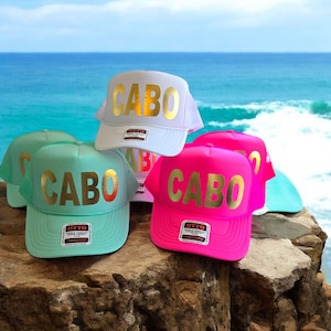 May include: Five trucker hats with mesh back panels. The hats are white, pink, and light blue. Each hat has the word "CABO" written in gold letters on the front.