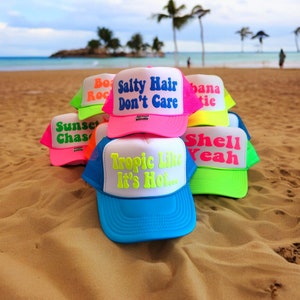 May include: A stack of six colorful trucker hats with neon and bright colored text. The hats have phrases like "Salty Hair Don't Care", "Shell Yeah", "Tropic Like It's Hot", "Sunset Chase", "Bo Roc", and "Banana Tie".