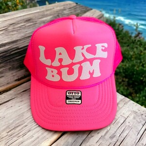 May include: A pink trucker hat with a white mesh back. The front of the hat has the words "LAKE BUM" in a retro font with silver glitter. The hat has a small black label that says "OTTO WORLD FAMOUS FOAM FRONT TRUCKER".