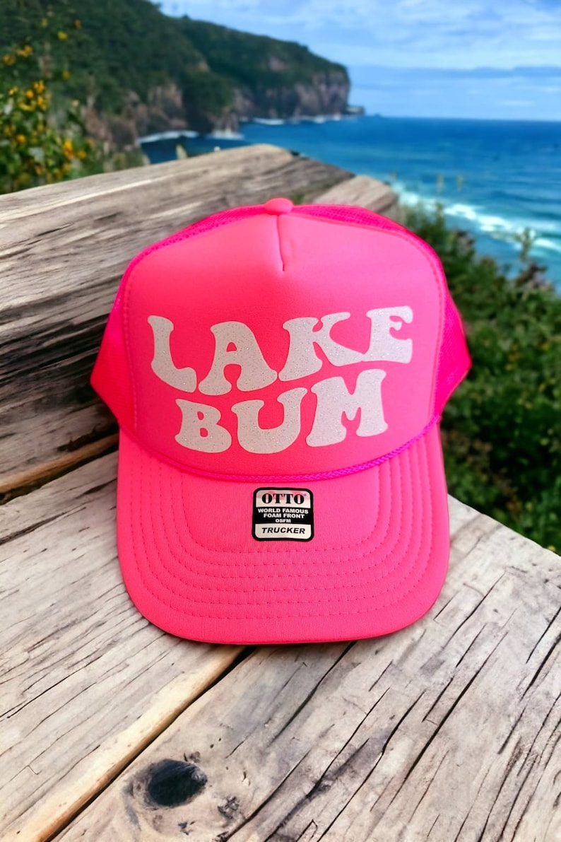 May include: A pink trucker hat with a white mesh back. The front of the hat has the words "LAKE BUM" in a retro font with silver glitter. The hat has a small black label that says "OTTO WORLD FAMOUS FOAM FRONT TRUCKER".