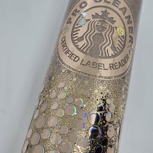 May include: A glittery tumbler with a gold and bronze design. The tumbler features a logo with the text "PRO CLEANER CERTIFIED LABEL READER". The design includes holographic bubble accents and a leopard print straw.