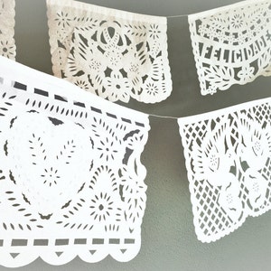 May include: White paper banner with cut-out designs, featuring the words "Felicidades" in Spanish. The banner is strung together with white string and hangs against a green wall.