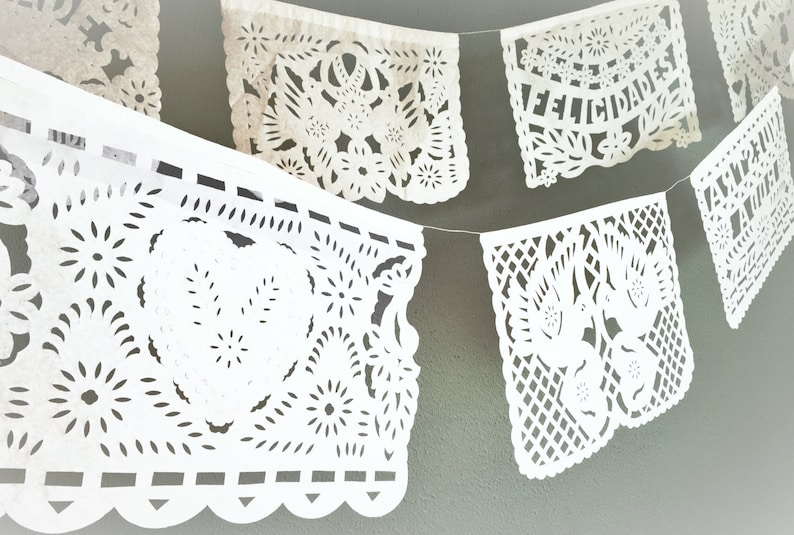 May include: White paper banner with cut-out designs, featuring the words "Felicidades" in Spanish. The banner is strung together with white string and hangs against a green wall.