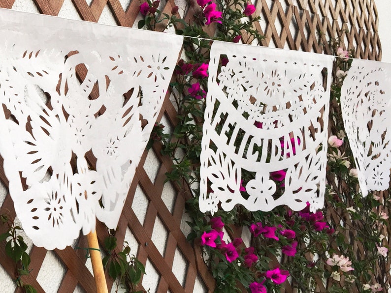 May include: White paper banners with cut-out designs, including flowers and the word "Felicidades", hanging against a wooden lattice background with pink flowers.