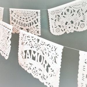 May include: White paper cut-out banners with the words "Felicidades" and "Amor" hanging on a string. The banners are decorated with intricate floral designs.