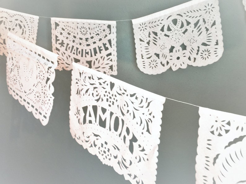 May include: White paper cut-out banners with the words "Felicidades" and "Amor" hanging on a string. The banners are decorated with intricate floral designs.