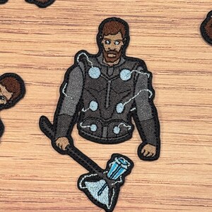 May include: Embroidered iron-on patch featuring a cartoon illustration of Thor from the Marvel Cinematic Universe. Thor is wearing his signature armor and holding his hammer, Mjolnir. The patch is black, gray, and blue.