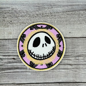 Könnte beinhalten: Ein gestickter Aufnäher mit dem ikonischen grinsenden Gesicht von Jack Skellington aus dem Film "Nightmare Before Christmas" in Pink und Gelb.