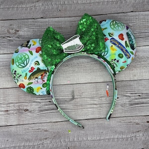 Könnte beinhalten: Grüne und blaue Minnie Mouse Ohren mit einer grünen Schleife und einem Aufdruck eines grünen Logos mit dem Text "Living with the Land" und einer Vielzahl von Früchten, Gemüse und Fischen.