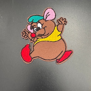 May include: An embroidered patch featuring a cartoon mouse wearing a green and blue hat, a yellow shirt, and red shoes. The mouse is walking and has a happy expression on its face.