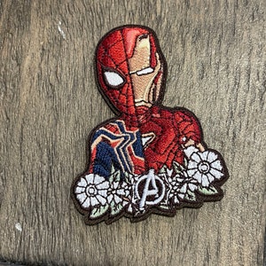 May include: Embroidered iron-on patch featuring a superhero with a red and blue costume, a white spider design, and a red and gold iron man mask. The superhero is surrounded by white flowers and a brown Avengers logo.