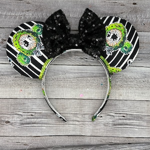 Pode incluir: Orelhas de rato listradas em preto e branco com um design de criatura verde e preta e um laço preto de lantejoulas.