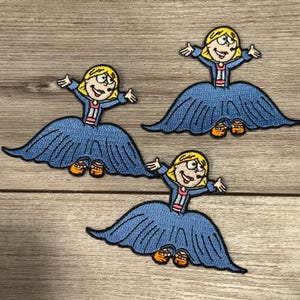 May include: Three embroidered patches featuring a cartoon girl with blonde hair, a blue dress, and orange shoes. The girl has her arms outstretched and is smiling. The patches are on a wooden surface.