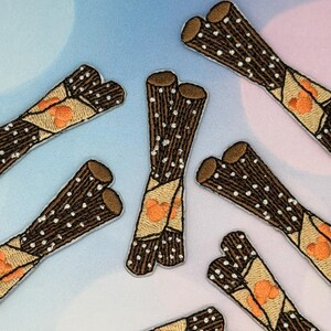 Könnte beinhalten: Gestickte Aufnäher mit einem braunen, zylindrischen Design mit weißen Tupfen und einer beigen Umhüllung mit orangefarbenen Akzenten. Die Aufnäher sind auf einem hellblauen und rosa Farbverlaufshintergrund angeordnet. Die Aufnäher scheinen ein Lebensmittelthema zu haben.