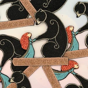 Może przedstawiać: Haftowane naszywki przedstawiające czarnego niedźwiedzia i rudowłosą postać w niebieskiej sukience. Niedźwiedź ma białe szwy i wchodzi w interakcję z postacią. Naszywki są na białym tle z brązowymi elementami dekoracyjnymi.