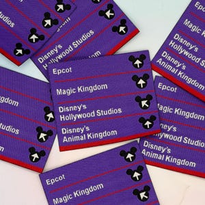 Puede incluir: Etiquetas de tela moradas con ribete rojo y texto blanco. Las etiquetas presentan el texto "Epcot", "Magic Kingdom", "Disney's Hollywood Studios" y "Disney's Animal Kingdom". Cada etiqueta tiene un icono de Mickey Mouse.