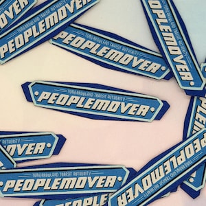 May include: Several blue and white fabric patches are displayed. Each patch features the text "PEOPLEMOVER" in white, with "TOMORROWLAND TRANSIT AUTHORITY" above. The patches are scattered on a light background.