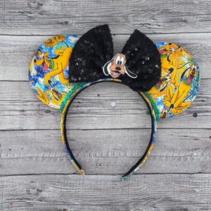 Può includere: Un paio di orecchie da topo nere e gialle con un fiocco nero di paillettes. Le orecchie presentano una stampa di Pluto il cane.