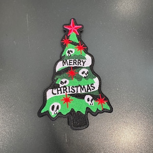 Könnte beinhalten: Grüne Weihnachtsbaum-Stickerei mit einem roten Stern oben, Totenköpfen und den Worten "Merry Christmas" in einem Banner.