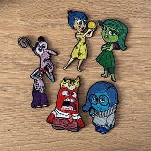 May include: Five embroidered patches featuring characters from the animated film Inside Out. The characters are Joy, Sadness, Anger, Fear, and Disgust. Each patch is colourful and detailed, with a unique expression on each character's face.