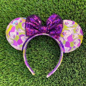 Pu&ograve; includere: Orecchie di Minnie Mouse viola e verdi con un fiocco di paillettes viola. Le orecchie sono ricoperte da una stampa di personaggi dei cartoni animati viola e verdi.