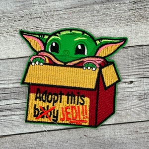 May include: Green and pink embroidered patch of a baby Yoda character peeking out of a red and yellow box with the text "Adopt this baby Jedi!!"