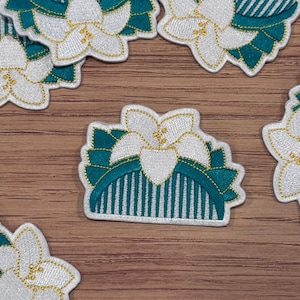 May include: A set of embroidered iron-on patches featuring white flowers with gold accents on a teal green background. The patches are in the shape of a flower and a half-circle.