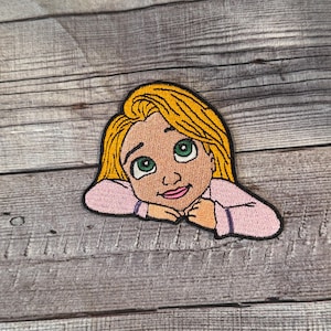 Op de afbeelding: Gestikt patch van een cartoonmeisje met lang blond haar, dat een roze shirt draagt en omhoog en naar rechts kijkt.