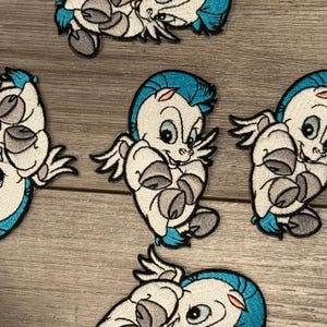 Könnte beinhalten: Mehrere gestickte Aufnäher mit einem Cartoon-Pegasus. Die Aufnäher haben weiße Körper, graue Hufe und türkisfarbene Akzente auf Mähne und Schwanz. Der Pegasus hat einen lächelnden Ausdruck und kleine Flügel. Die Aufnäher liegen auf einer hellbraunen Holzoberfläche.