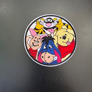 May include: Embroidered patch featuring Winnie the Pooh and friends: Tigger, Piglet, Eeyore, and Pooh Bear. The patch is circular with a white border and black outline. The characters are in vibrant colors, with a black background.