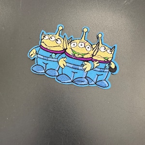 May include: Three embroidered alien characters from the Toy Story franchise. The aliens are green with three eyes and are wearing blue spacesuits. They are standing side by side with their arms around each other.