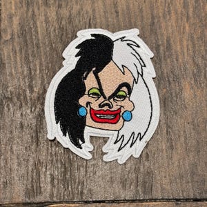 Może przedstawiać: Naszywka haftowana przedstawiająca Cruellę De Vil z filmu Disneya 101 dalmatyńczyków. Naszywka przedstawia jej twarz z przebiegłym uśmiechem, czarno-białe włosy i niebieskie kolczyki.