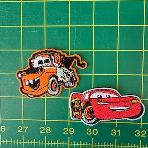 May include: Two embroidered iron-on patches featuring characters from the Disney Pixar movie Cars. The first patch is orange and depicts Mater, a tow truck with a wide grin. The second patch is red and depicts Lightning McQueen, a race car with a wide smile.