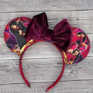 Könnte beinhalten: Stirnband mit burgunderfarbenem Samt-Schleife und roten und violetten Stoffohren mit einem Aufdruck einer Frau mit dunklen Haaren und einem roten Kleid. Die Ohren sind mit goldenen Pailletten verziert.