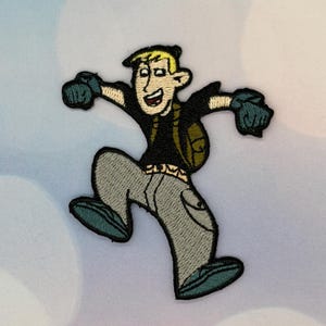 May include: Embroidered patch of a cartoon character in a leaping pose. The character has blonde hair, a black top, grey trousers, and a green backpack. The patch is detailed with teal accents and a smiling expression.
