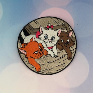 以下が含まれることがあります： オレンジ、白、茶色の3匹の漫画の猫が描かれた刺繍パッチ。猫は、テクスチャード加工された背景を持つ円形のデザインに配置されています。1匹の猫はピンクのリボンをしています。