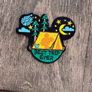 May include: A green, yellow, and black embroidered patch with a Mickey Mouse silhouette inside a tent. The text "BEST TRIP EVER" is written on the patch.