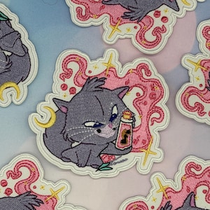 May include: An embroidered patch featuring a grey cat with a mischievous expression holding a small bottle with a silhouette design. The cat is surrounded by pink swirling smoke, yellow stars, and a crescent moon on a white background.