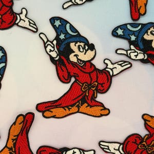 Może przedstawiać: Haftowane naszywki przedstawiające Myszkę Miki jako ucznia czarnoksiężnika z Fantazji. Naszywki przedstawiają Myszkę Miki w czerwonej szacie i niebieskim kapeluszu czarodzieja z gwiazdami. Naszywki są białe, czerwone, czarne i niebieskie.
