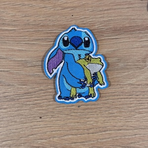 Könnte beinhalten: Gestickter Aufnäher mit einer blauen Cartoonfigur, die einen grünen Frosch hält. Die Figur hat große schwarze Augen und lila Ohren. Der Frosch ist gelb und grün. Der Aufnäher befindet sich auf einem hellblauen Hintergrund.