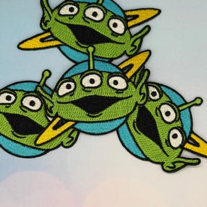 Op de afbeelding: Geborduurde patches met groene aliens met drie ogen, grote glimlachen en antennes. De aliens staan op blauwe cirkels met gele vleugelachtige accenten. De patches zijn waarschijnlijk voor kleding of accessoires.