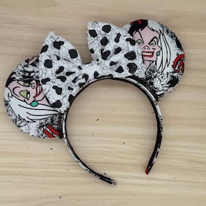 May include: A pair of black and white mouse ears with a sequined bow. The ears feature a Cruella de Vil design. The headband is black with a white and black pattern. The bow is white with black spots.