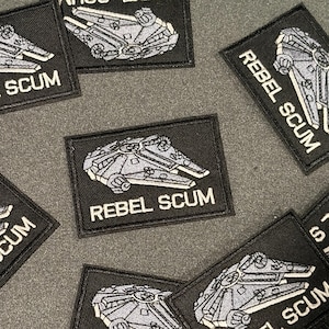 May include: Black rectangular patches featuring a gray spaceship design and the words "REBEL SCUM" in white. The patches are embroidered with a detailed image of the spaceship. The patches are scattered on a gray surface.