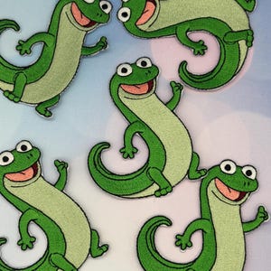 May include: Embroidered patches featuring a cartoon lizard in shades of green. The lizards have white eyes, pink mouths, and are in various poses. The patches are on a light blue and white background.