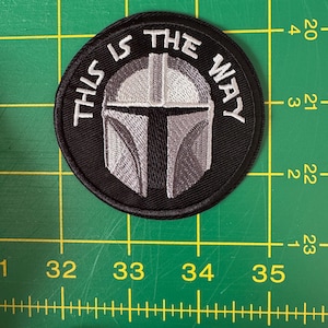 May include: A round, black embroidered patch with the text "THIS IS THE WAY" in white. The patch features a detailed, silver and black Mandalorian helmet design in the center. The patch is approximately 3 inches in diameter.