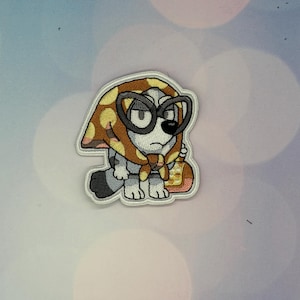 May include: An embroidered patch featuring a cartoon dog character wearing a brown and yellow polka-dot headscarf and large black-rimmed glasses. The character has grey and white fur, a black nose, and is holding a small yellow and pink bag.